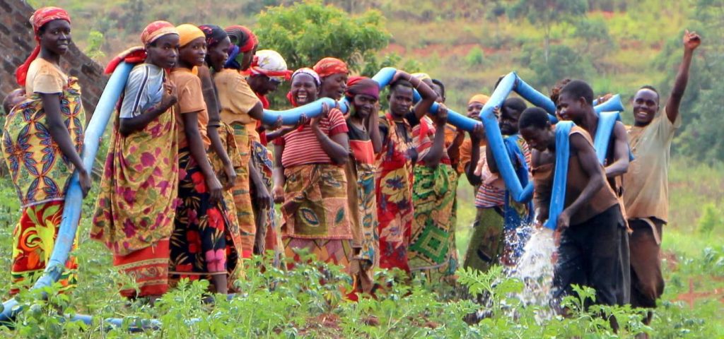 Group of people, mostly women in brightly colored clothes, holding long hose that is spewing water.
