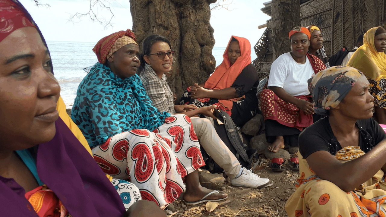 Small group of women, most in bright, patterned clothes, sitting outside, water in background.