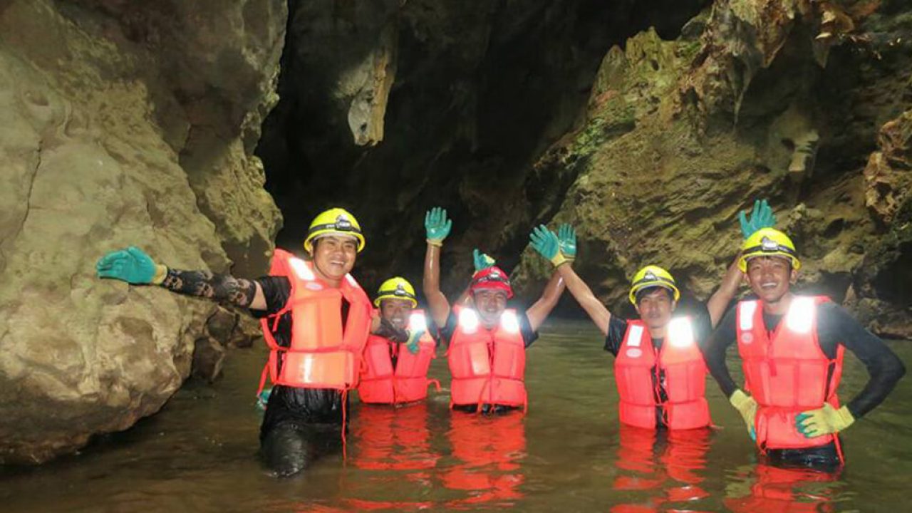 Five people wearing helmets and orange vests in cave, waist-height in water, smiling, waving at camera.