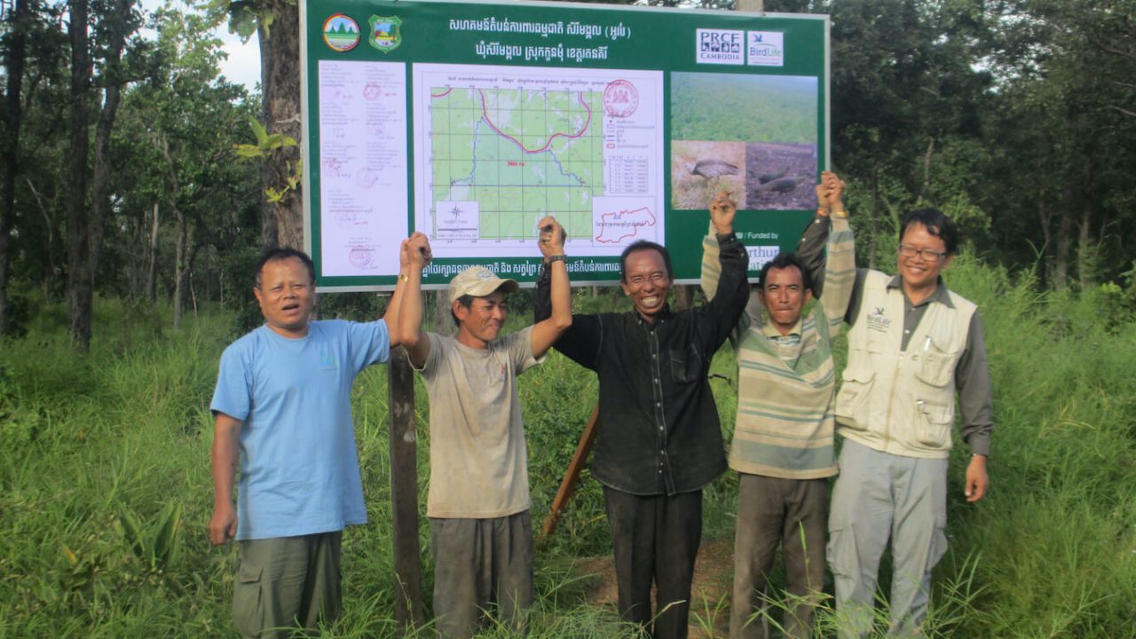 Group of 5 men standing outside, holding hands in the air and smiling in front of signboard with map.