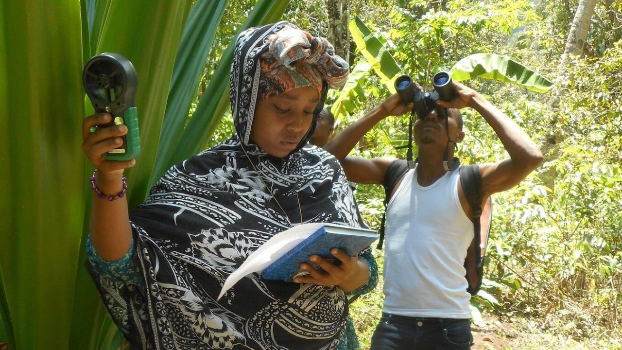 Woman looks at notebook while holding device up while man behind her looks through binoculars.