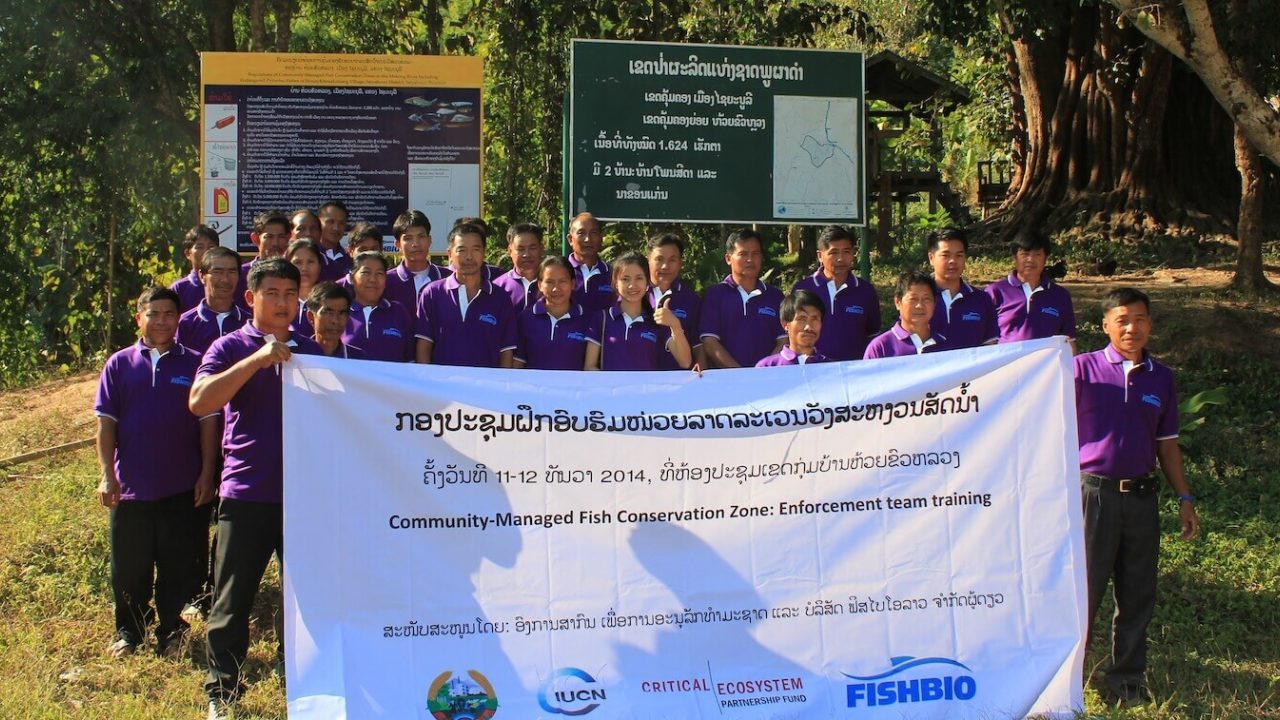 A group of about 25 men and women wearing purple shirts, standing behind signboard and holding large banner.