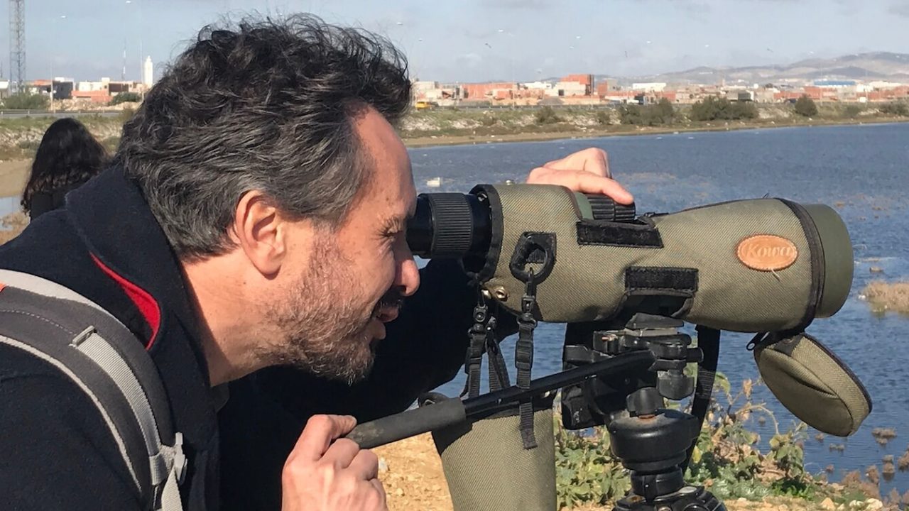 Close-up of man looking through a monocular, water behind him and buildings in background.