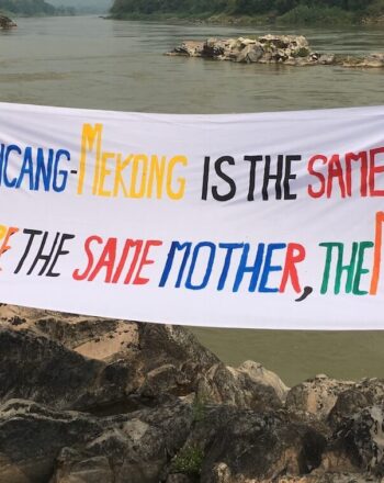 Two girls standing in front of river holding hand-made sign that says "Lancang-MEkong is the Same River We Share the Same Mother, the Mekong"