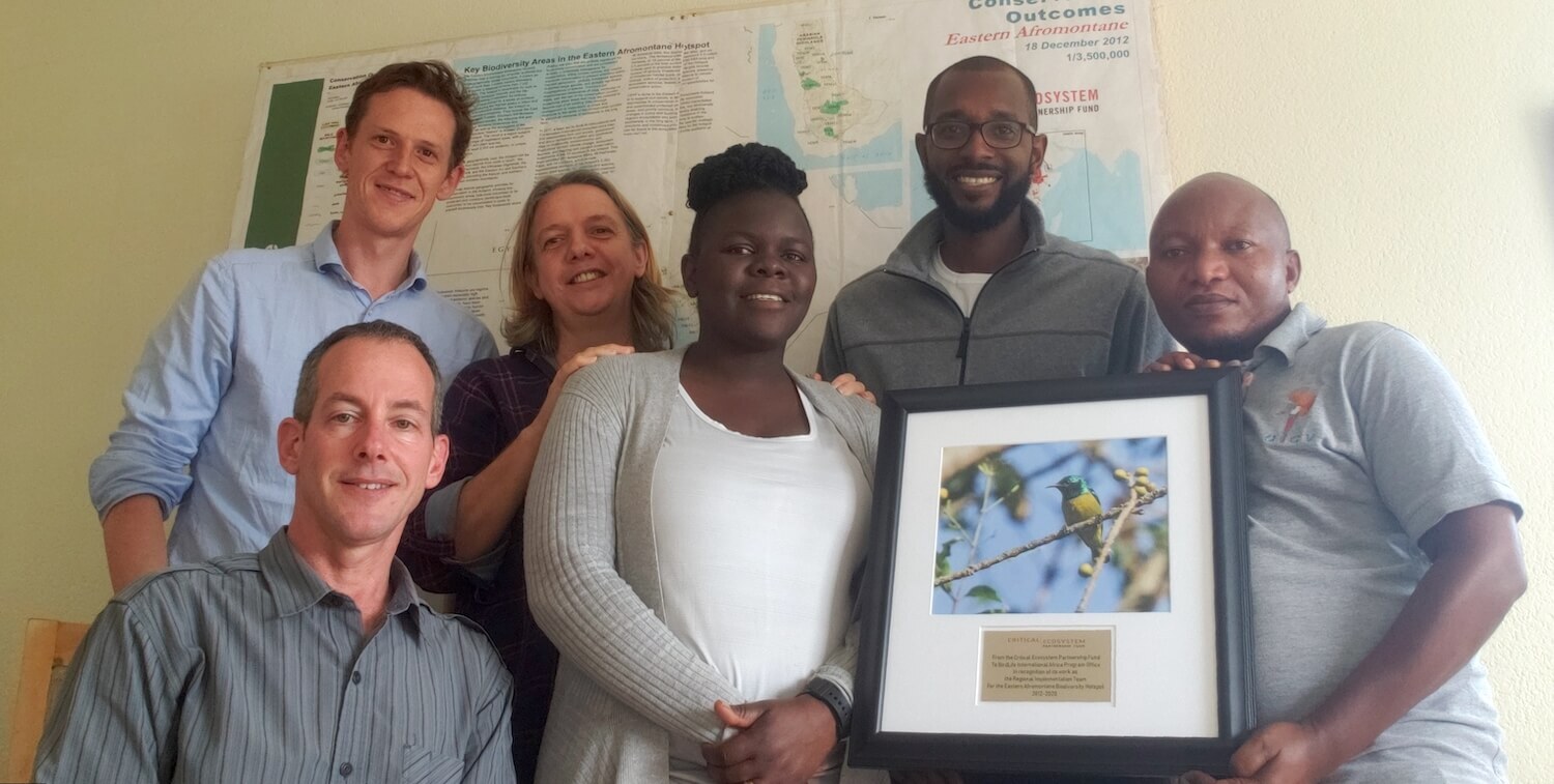 6 people indoors, smiling at camera. one person holds plaque with bird photo on it.