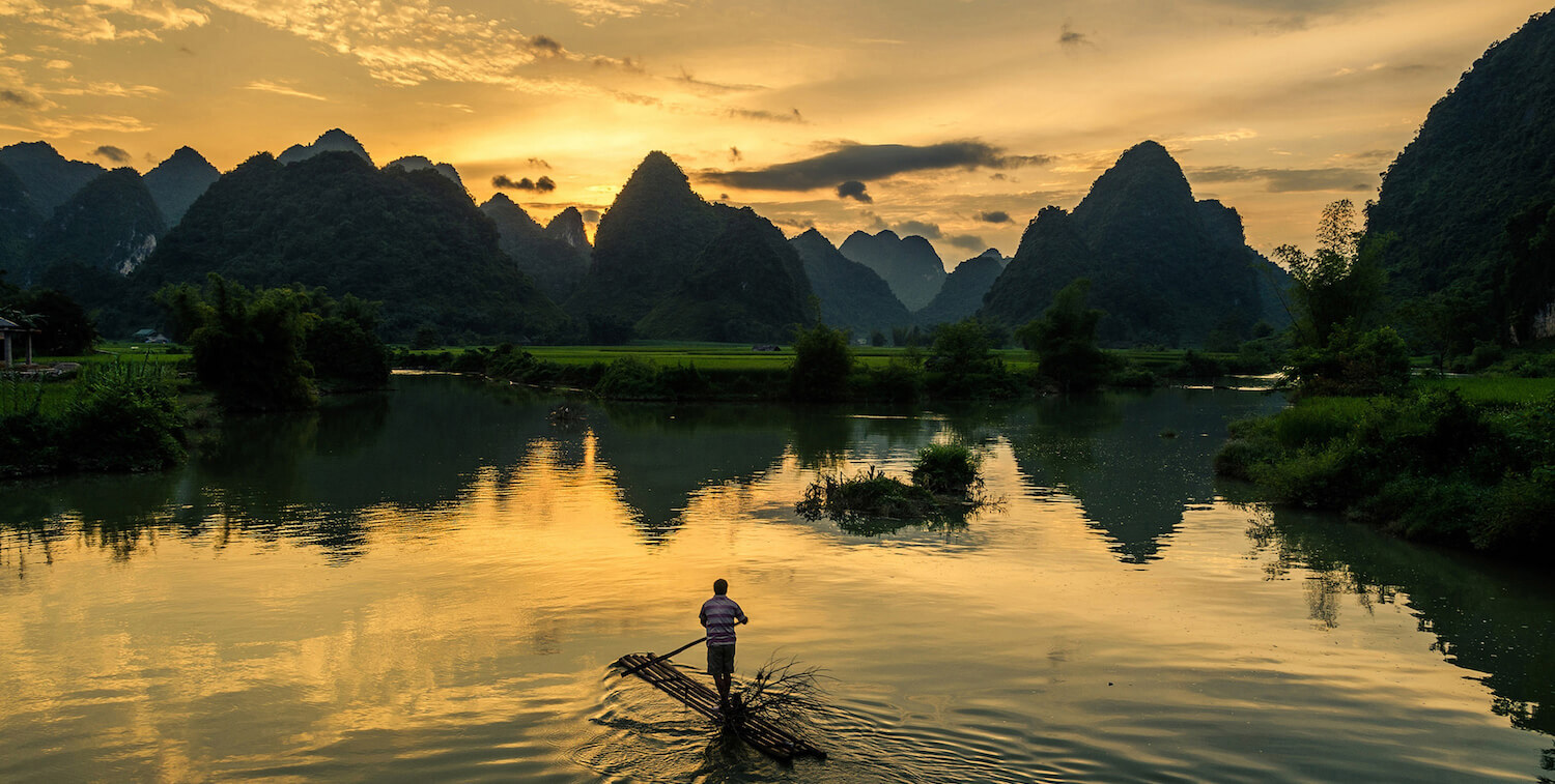 Man standing on thin raft made of logs, paddling across water as sun sets.