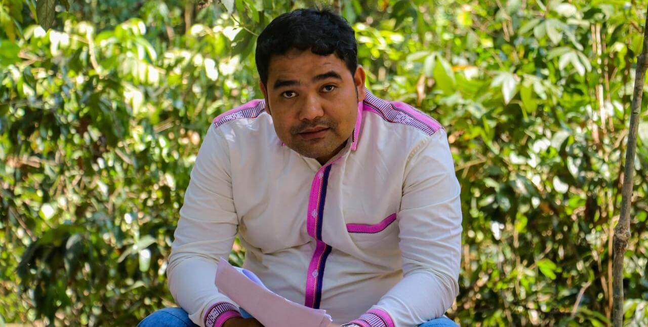 Man with white and pink shirt sitting outside, looking at camera.