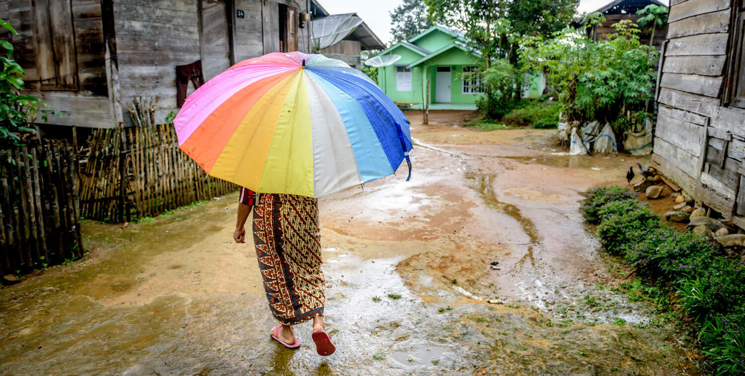 Woman with her back to camera holds a large, bright umbrella. She is walking on a dirt road, a few houses on either side.